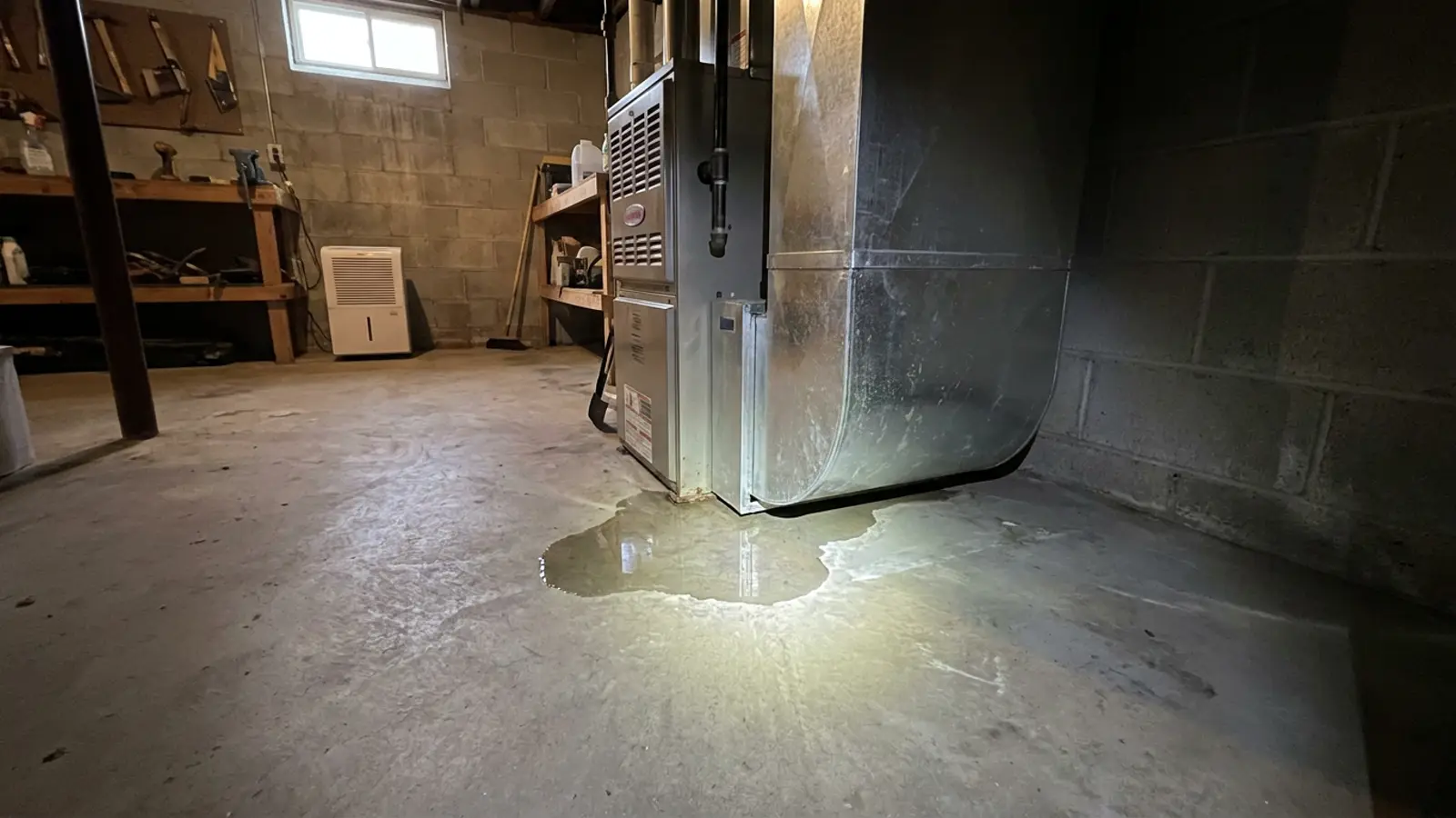 A small puddle of clear water spreading across a basement concrete floor beneath an indoor air handler