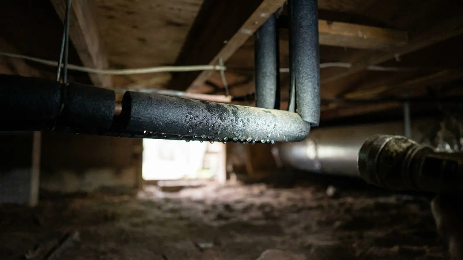 Condensation droplets clinging to the underside of insulated refrigerant lines in a dim crawlspace